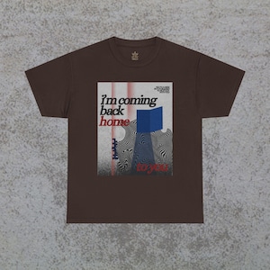 Puede incluir: Una camiseta marrón con un diseño gráfico que dice "I'm coming back home to you". El diseño incorpora un cubo azul y un patrón de remolino en blanco y negro. La camiseta tiene cuello redondo y mangas cortas.