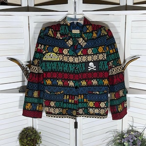 May include: A long-sleeve blazer featuring a vibrant, multi-colored woven pattern in red, blue, green, and yellow. The jacket has a notched lapel, two front pockets, and two black buttons. It includes a yellow patch and a black skull and crossbones patch.