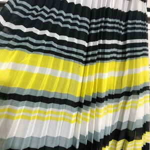May include: Pleated fabric with horizontal stripes in black, white, gray, and yellow. The fabric is folded in vertical pleats, creating a textured surface. The color scheme is bold and graphic.