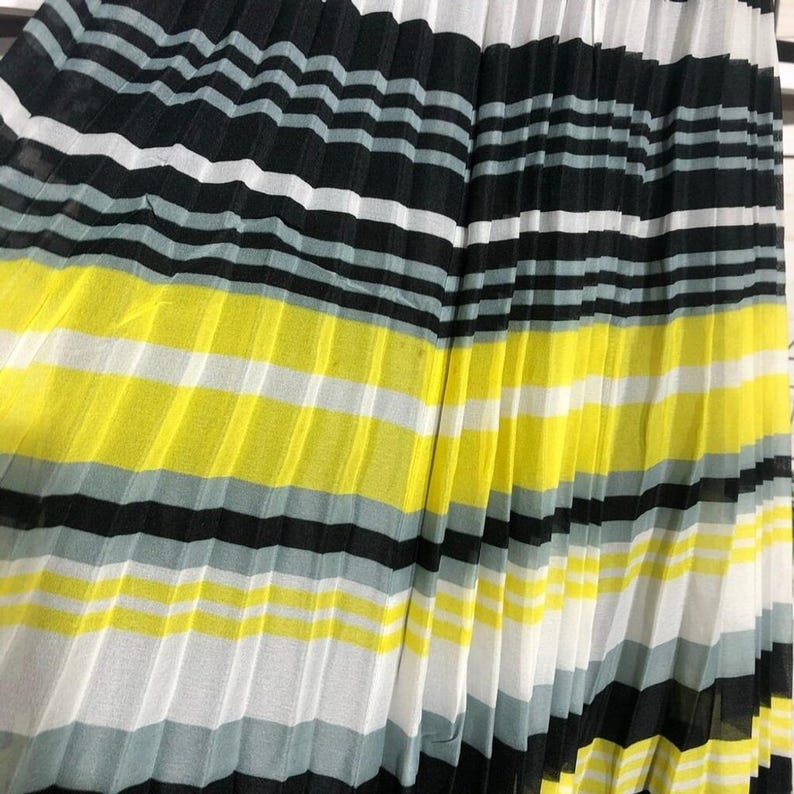 May include: Pleated fabric with horizontal stripes in black, white, gray, and yellow. The fabric is folded in vertical pleats, creating a textured surface. The color scheme is bold and graphic.
