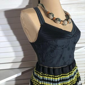 May include: A dark blue dress with a V-neck and floral embroidery on the bodice. The dress has a pleated skirt with yellow, black, and gray stripes. A beaded necklace with wooden and patterned beads is draped around the mannequin's neck.