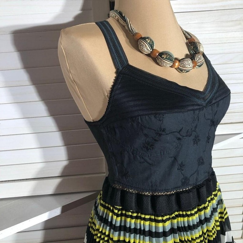 May include: A dark blue dress with a V-neck and floral embroidery on the bodice. The dress has a pleated skirt with yellow, black, and gray stripes. A beaded necklace with wooden and patterned beads is draped around the mannequin's neck.
