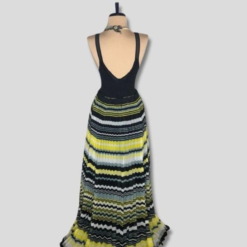 May include: A black sleeveless dress with a V-neck bodice and a pleated skirt. The skirt features horizontal stripes in yellow, black, white, and gray. The dress is displayed on a mannequin.