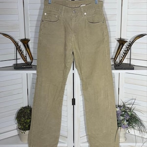 May include: Tan corduroy pants hanging on a metal hanger. The pants have a button and zipper closure, front and back pockets, and a straight leg design. The pants are made of a ribbed corduroy fabric.