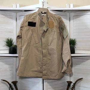 May include: Tan military-style jacket with a zip-up front, multiple pockets, and patches. The jacket has a high collar and long sleeves. A black rectangular patch and a tan shield-shaped patch are visible on the front.