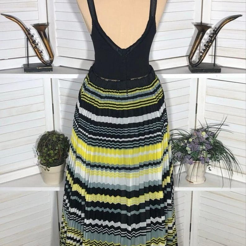 May include: A black sleeveless top with a V-neck is paired with a pleated skirt. The skirt features horizontal stripes in yellow, white, black, and gray. The dress is displayed on a mannequin.