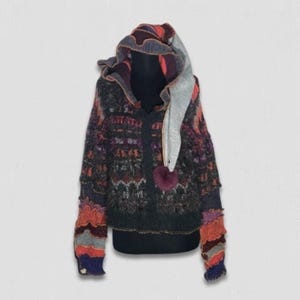 May include: A long-sleeve hooded sweater featuring a dark charcoal base with a multi-colored patterned knit design. The hood includes a light gray fabric lining and a dark purple pom-pom. The cuffs and hem feature layered, textured fabric strips.