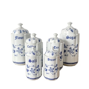 May include: A set of six white ceramic kitchen canisters with blue floral designs and matching lids. The canisters are labeled "Flour", "Coffee", "Salt", "Pepper", "Tea", and "Sugar". The canisters vary in size and shape.