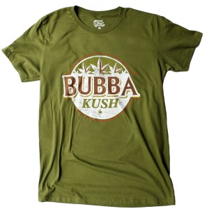 May include: An olive green t-shirt with a circular graphic. The graphic features the text "BUBBA KUSH" in brown and white, cannabis leaf illustrations, and a brown border. The shirt has a crew neck and short sleeves.