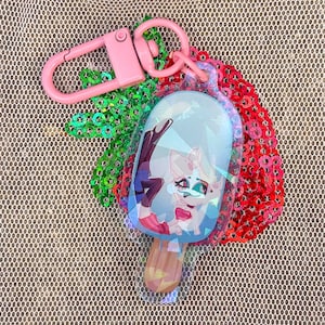 May include: A pink and white popsicle keychain with a cartoon character on it. The keychain is made of clear plastic and has a holographic finish. The character is a white fox with pink ears and a red bow. The keychain is attached to a pink metal ring.