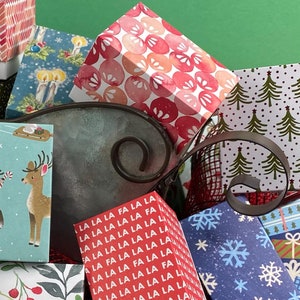 May include: A galvanized metal sleigh filled with various small gift boxes. The boxes feature festive patterns like Christmas lights, reindeer, and botanical designs. The background is a solid green, with a red and black mesh in the foreground.