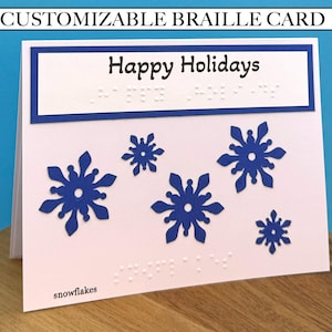 May include: A white greeting card featuring the text Happy Holidays in black ink with corresponding Braille dots below. The card is decorated with five blue paper snowflakes of varying sizes and the word snowflakes printed in black.