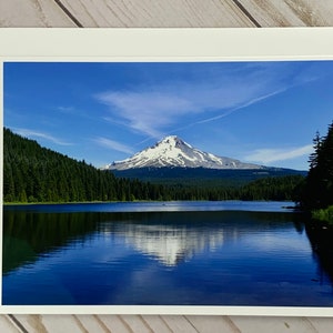 May include: A scenic photograph of a snow-capped mountain reflected in a still lake. The mountain is surrounded by a forest of evergreen trees. The sky is blue with white clouds.