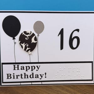 May include: A white birthday card with black accents. The card features three balloons, the number 16, and the words "Happy Birthday!" in a bold font. Braille dots are also present on the card.