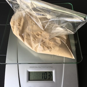 May include: A digital kitchen scale with a clear plastic bag containing a brown powder. The scale reads 103 grams. The scale has a maximum weight capacity of 5 kilograms or 11 pounds.