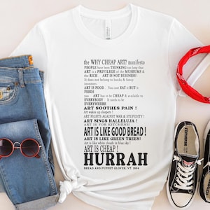 Puede incluir: Camiseta blanca con un manifiesto sobre arte, combinada con vaqueros azules, gafas de sol rojas, una bandana roja y zapatillas Converse negras. El texto de la camiseta dice "ART IS LIKE GOOD BREAD!" y "ART IS CHEAP!"