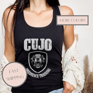 May include: Black tank top with the word "CUJO" in distressed white lettering. Below the word is a shield graphic with a snarling dog and the words "OBEDIENCE TRAINING". The tank top is worn by a person.
