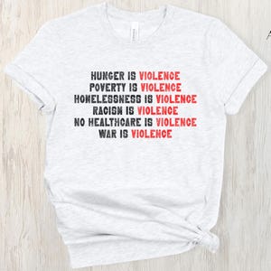 May include: Pale grey t-shirt featuring the statement "HUNGER IS VIOLENCE, POVERTY IS VIOLENCE, HOMELESSNESS IS VIOLENCE, RACISM IS VIOLENCE, NO HEALTHCARE IS VIOLENCE, WAR IS VIOLENCE" in black and red. The shirt is made of a soft material.