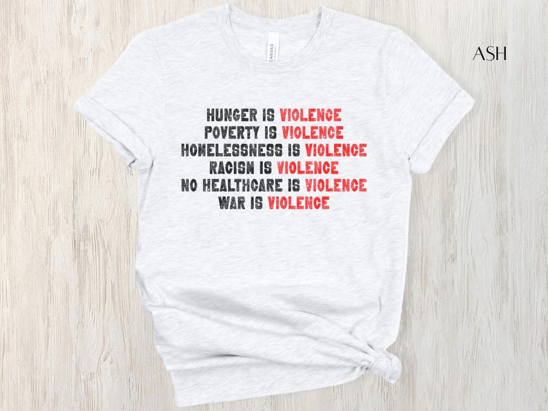May include: Pale grey t-shirt featuring the statement "HUNGER IS VIOLENCE, POVERTY IS VIOLENCE, HOMELESSNESS IS VIOLENCE, RACISM IS VIOLENCE, NO HEALTHCARE IS VIOLENCE, WAR IS VIOLENCE" in black and red. The shirt is made of a soft material.