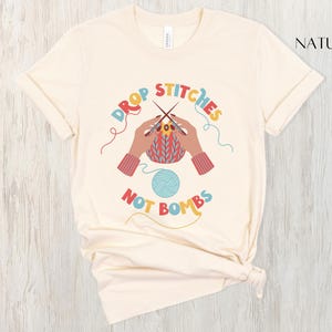 May include: Cream-colored t-shirt with the text "DROP STITCHES NOT BOMBS" in colorful lettering. The design features hands knitting, a ball of yarn, and knitting needles. The shirt is made of a soft material.