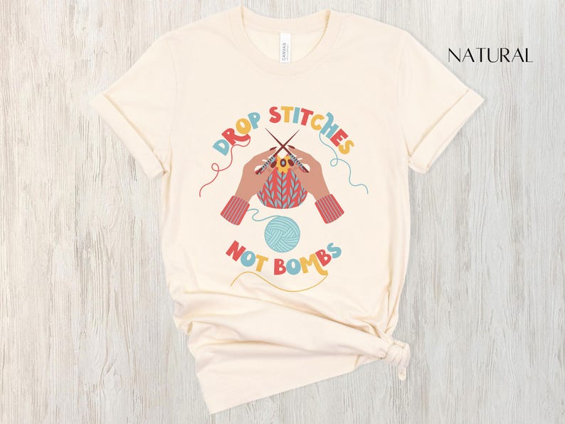 May include: Cream-colored t-shirt with the text "DROP STITCHES NOT BOMBS" in colorful lettering. The design features hands knitting, a ball of yarn, and knitting needles. The shirt is made of a soft material.