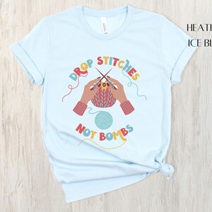 May include: Light blue t-shirt with the text "DROP STITCHES NOT BOMBS" in colorful letters. The graphic shows hands knitting with needles and yarn. The shirt is a heather ice blue color.
