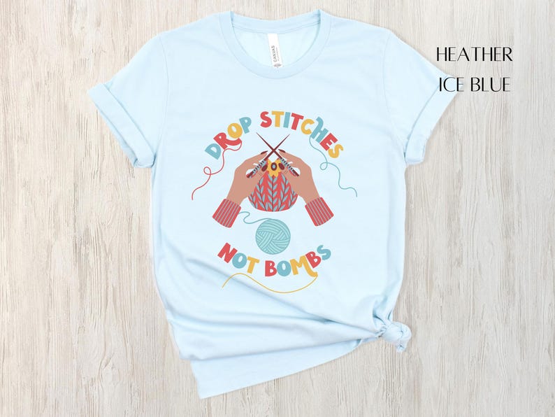 May include: Light blue t-shirt with the text "DROP STITCHES NOT BOMBS" in colorful letters. The graphic shows hands knitting with needles and yarn. The shirt is a heather ice blue color.