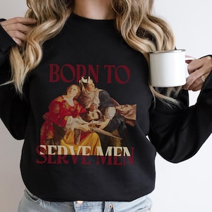 Op de afbeelding: Zwart sweatshirt met een schilderij van drie mensen en de tekst "Born to Serve Men".