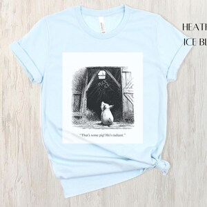 Puede incluir: Camiseta azul claro con una ilustraci&oacute;n en blanco y negro de un cerdo mirando hacia un granero. La ilustraci&oacute;n incluye el texto "That's some pig! He's radiant."