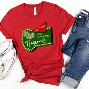 Può includere: Maglietta rossa con un cartello verde e bianco che dice "Fine Italian Steak & Seafood Truffoni's". Il cartello ha un cuoco dei cartoni animati e lampadine intorno al bordo.