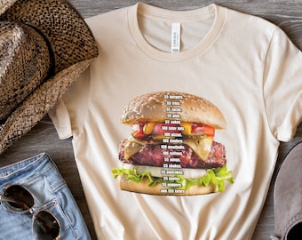 May include: A cream-colored crew neck t-shirt featuring a graphic of a cheeseburger with a list of food items printed over it, including 55 burgers, 55 fries, 55 tacos, 55 pies, 55 cokes, 100 tater tots, 100 pizzas, 100 tenders, 100 meatballs, 100 coffees, 55 wings, 55 shakes, 55 pancakes, 55 pastas, 55 peppers, and 155 taters.