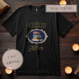 May include: Black t-shirt featuring a design with the text "The Manhattan Restaurant of the Mind". The design includes a stack of books within an ornate frame. The shirt is displayed on a dark wooden surface.