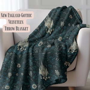 May include: A dark teal throw blanket with a floral and architectural pattern, draped over a white armchair. The blanket features a repeating design of white buildings and flowers. Text on the image reads "New England Gothic Velveteen Throw Blanket."