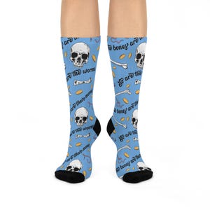 May include: Light blue crew socks featuring a repeating pattern of white skulls, bones, pink worms, and gold coins. The text "the bones are their money" and "so are the worms" is printed in black script across the fabric with black toe and heel sections.