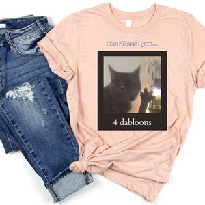 Peut inclure: Un t-shirt rose avec une image de chat noir et le texte "That'll cost you... 4 dabloons".