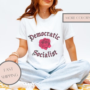 May include: A white short-sleeved t-shirt featuring the text Democratic Socialist in a dark gothic font with a pink rose graphic centered between the words. The shirt has a crew neckline and is worn with light blue denim jeans.