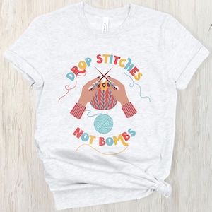 May include: Light gray t-shirt with the text "DROP STITCHES NOT BOMBS" in colorful letters. The graphic shows hands knitting with needles and yarn, and a ball of yarn. The shirt is a crew neck.