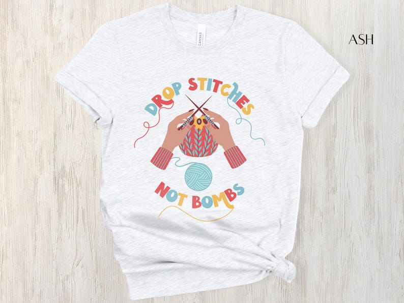 May include: Light gray t-shirt with the text "DROP STITCHES NOT BOMBS" in colorful letters. The graphic shows hands knitting with needles and yarn, and a ball of yarn. The shirt is a crew neck.