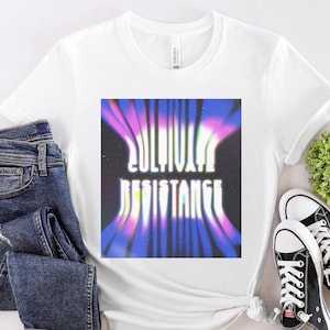 May include: White t-shirt featuring the text "CULTIVATE RESISTANCE" in a retro, wavy font. The graphic is a blend of purple, pink, and blue hues. The image also shows blue denim jeans and black Converse trainers.