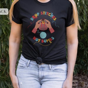 May include: Black t-shirt with the text "DROP STITCHES NOT BOMBS" in colorful lettering. The graphic features hands knitting with a ball of yarn. The shirt is worn by a person in blue jeans.