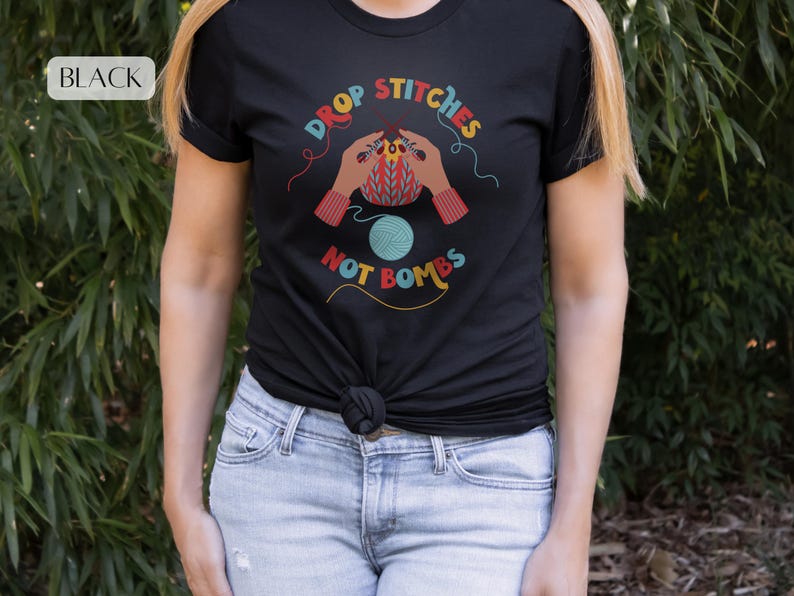 May include: Black t-shirt with the text "DROP STITCHES NOT BOMBS" in colorful lettering. The graphic features hands knitting with a ball of yarn. The shirt is worn by a person in blue jeans.