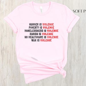 May include: Pale pink t-shirt featuring the statement "HUNGER IS VIOLENCE, POVERTY IS VIOLENCE, HOMELESSNESS IS VIOLENCE, RACISM IS VIOLENCE, NO HEALTHCARE IS VIOLENCE, WAR IS VIOLENCE" in black and red text.