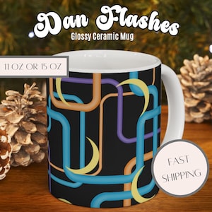 Può includere: Una tazza in ceramica lucida con il testo Dan Flashes e Glossy Ceramic Mug. La tazza presenta uno sfondo nero con un motivo di linee rettangolari arrotondate interconnesse in blu, arancione e viola, accentuato da forme a mezzaluna gialle.