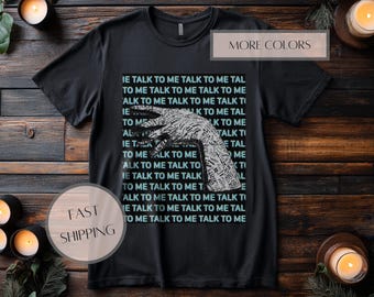 May include: A black short-sleeve t-shirt featuring a central graphic of a hand silhouette filled with the repeated text TALK TO ME. The background of the graphic consists of a grid of the same text in light blue, creating a layered typographic design.