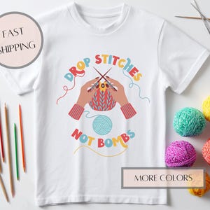May include: White t-shirt with a colorful graphic of hands knitting, the text "DROP STITCHES NOT BOMBS", and yarn balls. Knitting needles and pencils are also visible. The shirt is a craft-themed item.