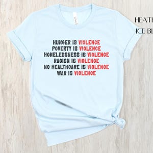 May include: Pale blue t-shirt with the statement "HUNGER IS VIOLENCE, POVERTY IS VIOLENCE, HOMELESSNESS IS VIOLENCE, RACISM IS VIOLENCE, NO HEALTHCARE IS VIOLENCE, WAR IS VIOLENCE" in black and red. The shirt is labelled "HEATHER ICE BLUE".