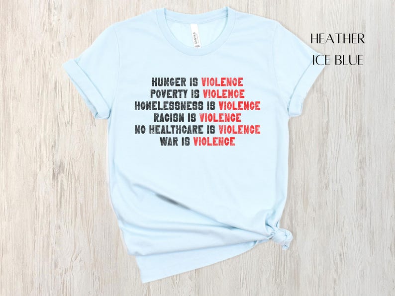 May include: Pale blue t-shirt with the statement "HUNGER IS VIOLENCE, POVERTY IS VIOLENCE, HOMELESSNESS IS VIOLENCE, RACISM IS VIOLENCE, NO HEALTHCARE IS VIOLENCE, WAR IS VIOLENCE" in black and red. The shirt is labelled "HEATHER ICE BLUE".