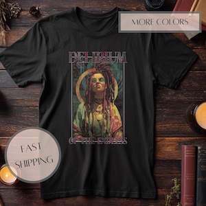 Op de afbeelding: Zwart t-shirt met een kleurrijke afbeelding van een persoon met dreadlocks en de tekst "DELIRIUM OF THE ENDLESS". Het ontwerp bevat een gouden halo en sierlijke sieraden. Het shirt wordt getoond op een donker houten oppervlak.