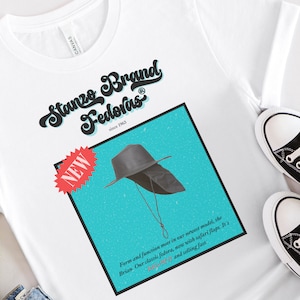 Puede incluir: Una camiseta blanca con un gráfico azul y blanco de un sombrero fedora negro. El gráfico incluye el texto "Stanze Brand Fedoras since 1963" y "NEW".