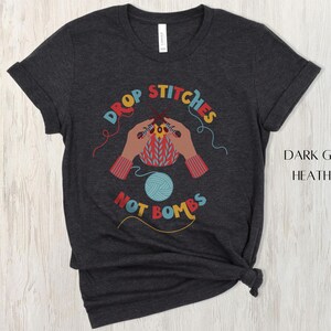 May include: Dark grey heather t-shirt with a colorful graphic. The graphic features hands knitting with the text "DROP STITCHES NOT BOMBS" in a circular design. A ball of blue yarn is also in the design.